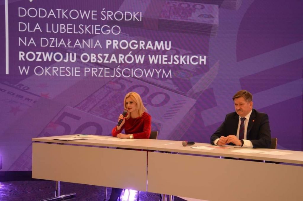 Zdjęcie przedstawiające dwie osoby, kobietę i mężczyznę siedzących przy stole konferencyjnym i udzielających wywiadu. Za nimi w tle znajduje się ogromny telebim z napisem Dodatkowe środki dla lubelskiego na działania Programu Rozwoju Obszarów wiejskich w okresie przejściowym Ponad 100 mln euro dla województwa lubelskiego na realizację Programu Rozwoju Obszarów Wiejskich na lata 2014-2020