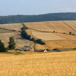 Obrazek przedstawia pola uprawowe z przewagą zbóż. W środku zdjęcia zabudowania oraz maszyny rolnicze.