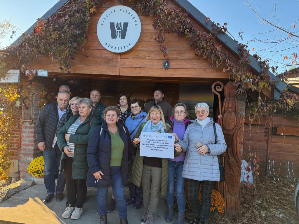 Grupa uczestników stojąca przed drewnianym budynkiem z szyldem Winnica Widokowa Komboria