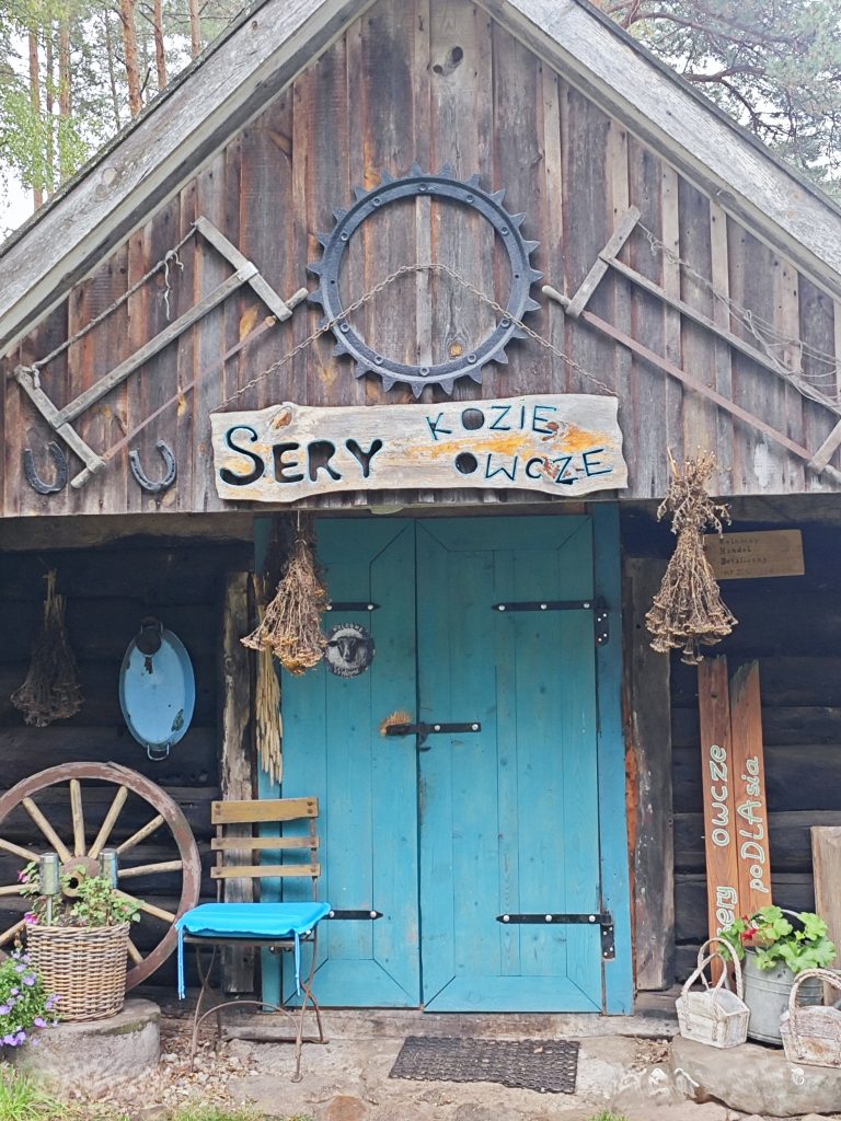 Drewniany domek w którym jest serowarnia. Stary drewniany domek z niebieskimi drzwiami z drewnianym szyldem „Sery owcze i kozie” ozdobiony jest podkowami, starymi narzędziami, bukietami suszonych kwiatów. Oparte jest o jego frontową ścianę koło od wozu, stoi niebieskie drewniane krzesło, wiklinowe kosze. Wszystko w sielskim folkowym stylu.