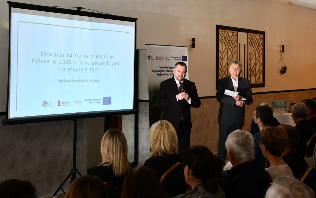 Dwóch mężczyzn ubranych elegancko, stoją przy ekranie na którym jest wyświetlany napis Sytuacja na rynku chmielu w Polsce w 2025 roku oraz perspektywa na przyszłe lata. Kilka siedzących osób.
