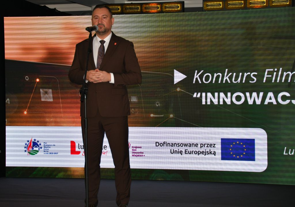 Marek Wojciechowski - Wicemarszałek Województwa Lubelskiego ubrany elegancko stoi na scenie, przy mikrofonie na statywie. W tle wyświetla się kolorowa grafika z logotypami Unii Europejskiej, Planu Strategicznego dla Wspólnej Polityki Rolnej na lata 2023-2027, oraz napis Dofinansowane przez Unię Europejską.