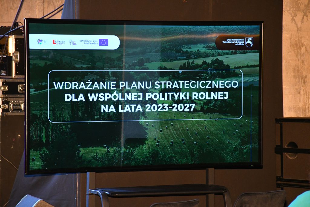 Grafika informująca o konferencji „Wdrażanie Planu Strategicznego dla Wspólnej Polityki Rolnej na lata 2023 – 2027”. Napis widnieje na tle zdjęcia pola na którym leżą bele siana i użytków zielonych, w dali widać las. Wszystko jest w tonacji ciemnozielonej.