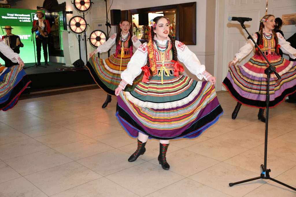 Trzy kobiety, śpiewają, ubrane w kolorowe stroje ludowe. W tle scena , na której siedzi mężczyzna ubrany w strój ludowy, gra na akordeonie.