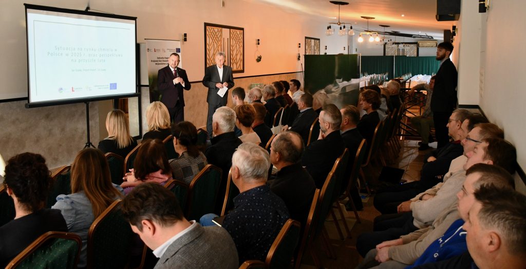 Grupa osób siedząca na krzesłach i słuchająca przemowy mężczyzny ubranego elegancko.