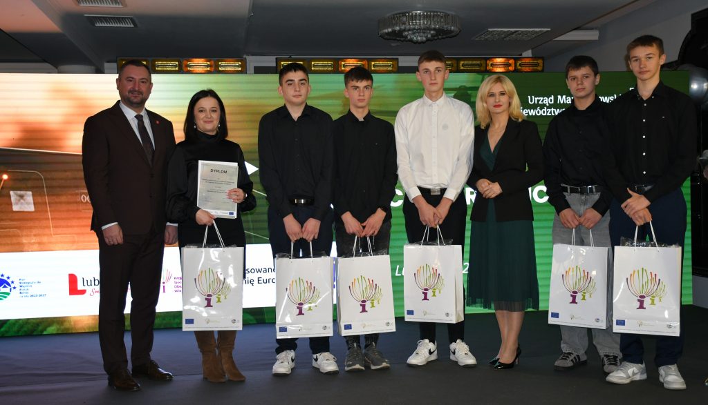 2. Na scenie, na tle kolorowej grafiki stoi grupa osób (dwie kobiety i sześciu mężczyzn) ubranych elegancko, w dłoniach trzymają białe torby prezentowe.