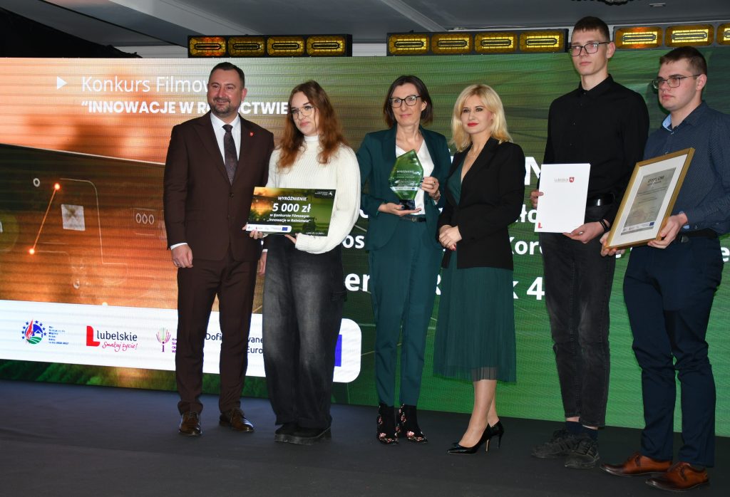 Na scenie, na tle kolorowej grafiki stoi grupa osób (trzy kobiety i trzech mężczyzn) ubranych elegancko, w dłoniach trzymają czek, statuetkę, dyplom i białą teczkę.