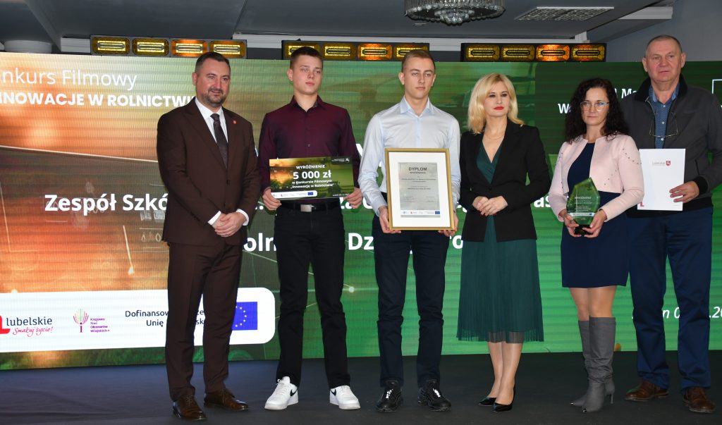 Na scenie, na tle kolorowej grafiki stoi grupa osób (dwie kobiety i czterech mężczyzn) ubranych elegancko, w dłoniach trzymają czek, statuetkę, dyplom i białą teczkę.