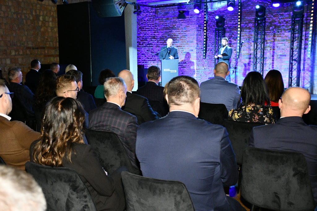 Marszałek województwa lubelskiego Jarosław Stawiarski stoi na scenie przy mównicy i mówi do uczestników konferencji „Wdrażanie Planu Strategicznego dla Wspólnej Polityki Rolnej na lata 2023 – 2027”. Obok niego stoi prowadzący konferencję.