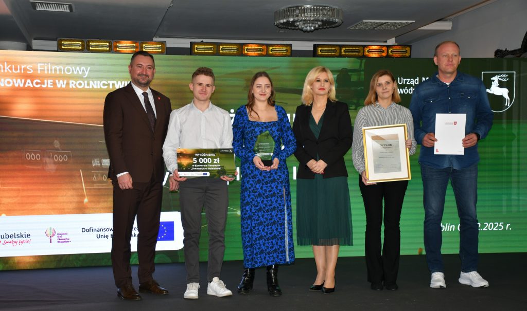 Na scenie, na tle kolorowej grafiki stoi grupa osób (trzy kobiety i trzech mężczyzn) ubranych elegancko, w dłoniach trzymają czek, statuetkę, dyplom i białą teczkę.