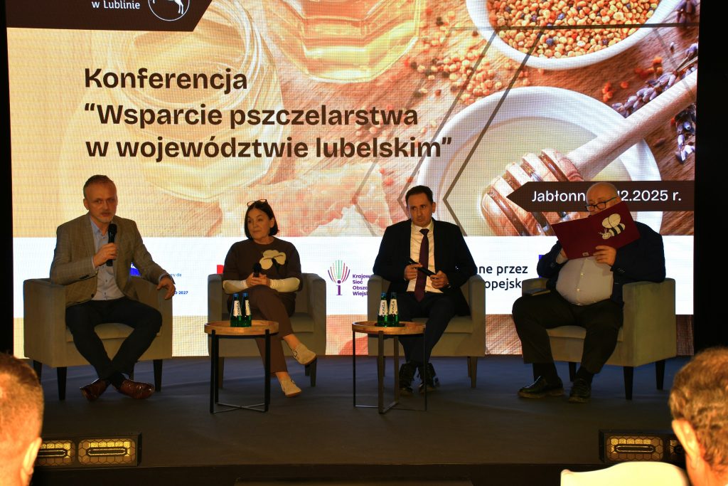 Na scenie na fotelach siedzą cztery osoby. Są to prelegenci oraz moderator, który prowadzi panel dyskusyjny podczas konferencji „Wsparcie pszczelarstwa w województwie lubelskim”.