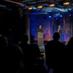 Marszałek województwa lubelskiego Jarosław Stawiarski stoi na scenie przy mównicy i mówi do uczestników konferencji „Wdrażanie Planu Strategicznego dla Wspólnej Polityki Rolnej na lata 2023 – 2027”. Obok niego stoi prowadzący konferencję.