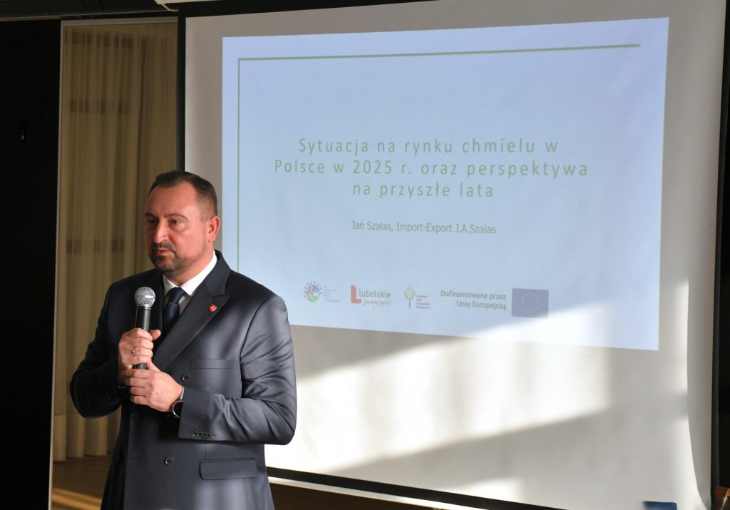 Mężczyzna ubrany elegancko, stoi na tle ekranu na którym wyświetla się napis Sytuacja na rynku chmielu w Polsce w 2025 roku oraz perspektywa na przyszłe lata, zestaw logotypów, znaczek Unii Europejskiej, napis Lubelskie Smakuj Życie, Znaczek KSOW+ oraz znaczek PS WPR 2023-2027 oraz napis Dofinansowane przez Unię Europejską.