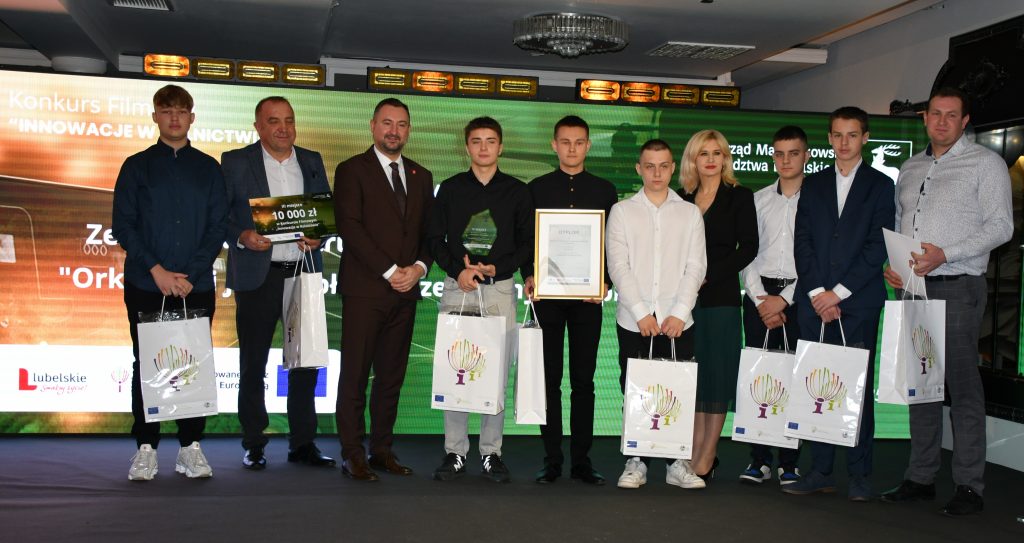 Na scenie, na tle kolorowej grafiki stoi grupa osób (kobieta i dziewięciu mężczyzn) ubranych elegancko, w dłoniach trzymają czek, statuetkę, dyplom i białą teczkę oraz białe torby prezentowe.