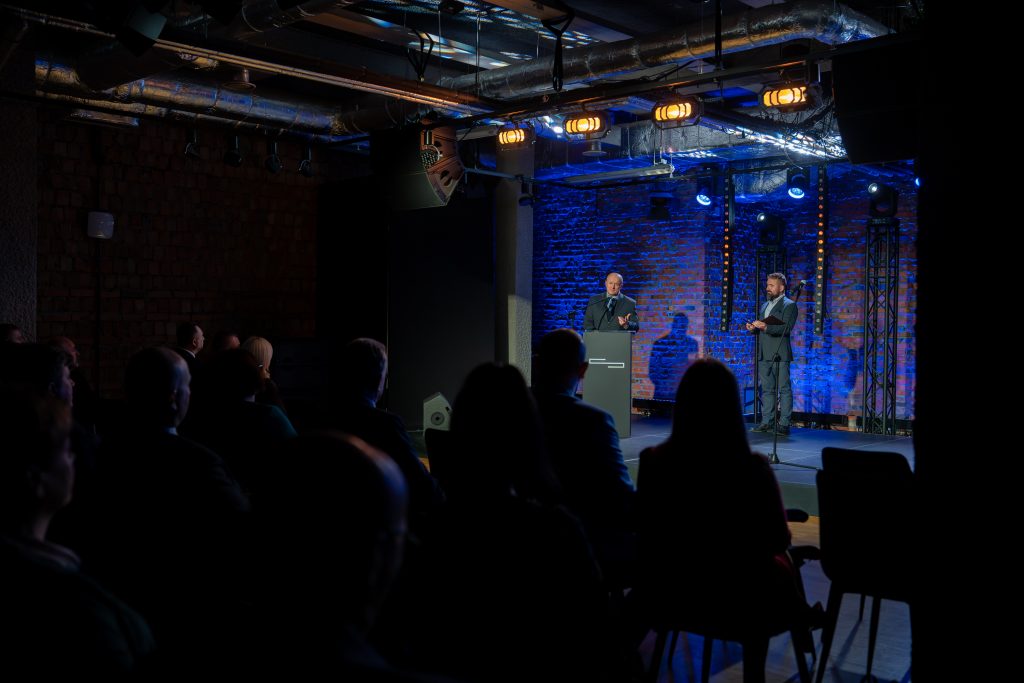 6. Marszałek województwa lubelskiego Jarosław Stawiarski stoi na scenie przy mównicy i mówi do uczestników konferencji „Wdrażanie Planu Strategicznego dla Wspólnej Polityki Rolnej na lata 2023 – 2027”. Obok niego stoi prowadzący konferencję.