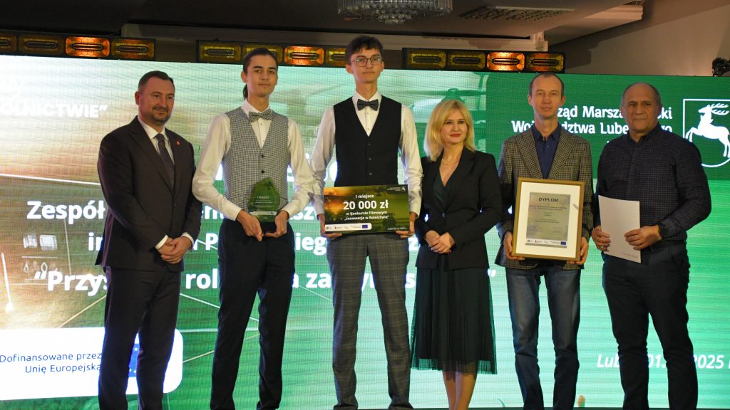 Na scenie, na tle kolorowej grafiki stoi grupa osób (kobieta i pięciu mężczyzn) ubranych elegancko, w dłoniach trzymają czek, statuetkę, dyplom i białą teczkę.