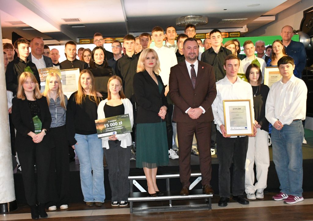 Na scenie, na tle kolorowej grafiki stoi grupa osób ubranych elegancko, w dłoniach trzymają czeki, statuetki, dyplomy.