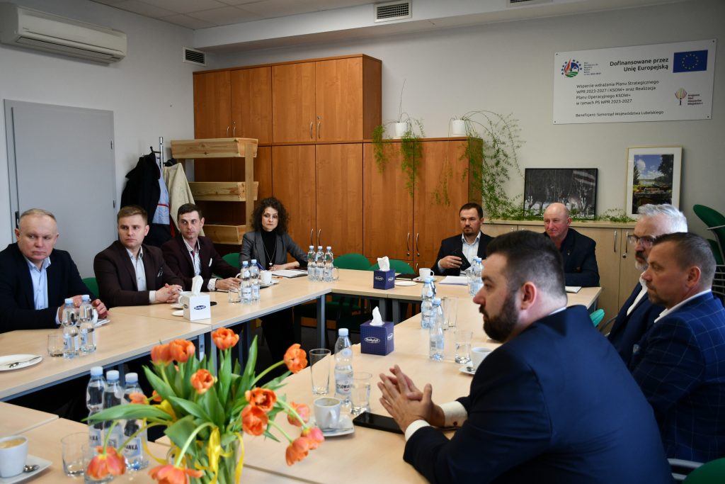 Mężczyźni oraz kobieta ładnie ubrani, siedzący przy stołach ustawionych na przeciwko siebie.