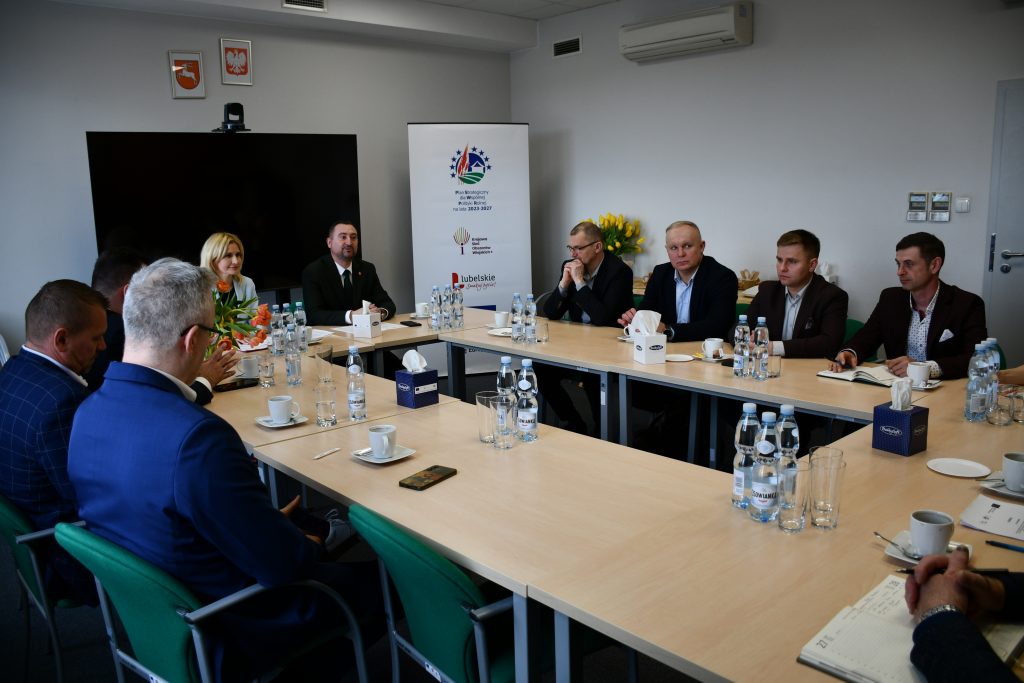 Mężczyźni oraz kobieta ładnie ubrani, siedzący przy stołach ustawionych na przeciwko siebie.