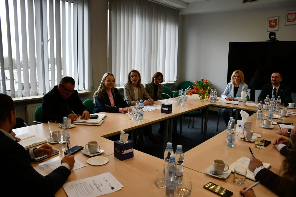 Mężczyźni oraz kobiety ładnie ubrani, siedzący przy stołach ustawionych na przeciwko siebie.