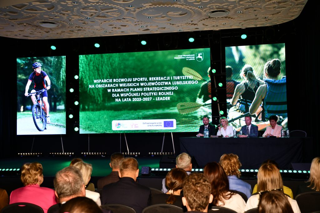 Uczestnicy konferencji „Wsparcie rozwoju sportu, rekreacji i turystyki na obszarach wiejskich województwa lubelskiego w ramach Planu Strategicznego dla Wspólnej Polityki Rolnej na lata 2023–2027 – LEADER” siedzą na Sali konferencyjnej i oglądają prezentację przedstawiającą dobre praktyki.