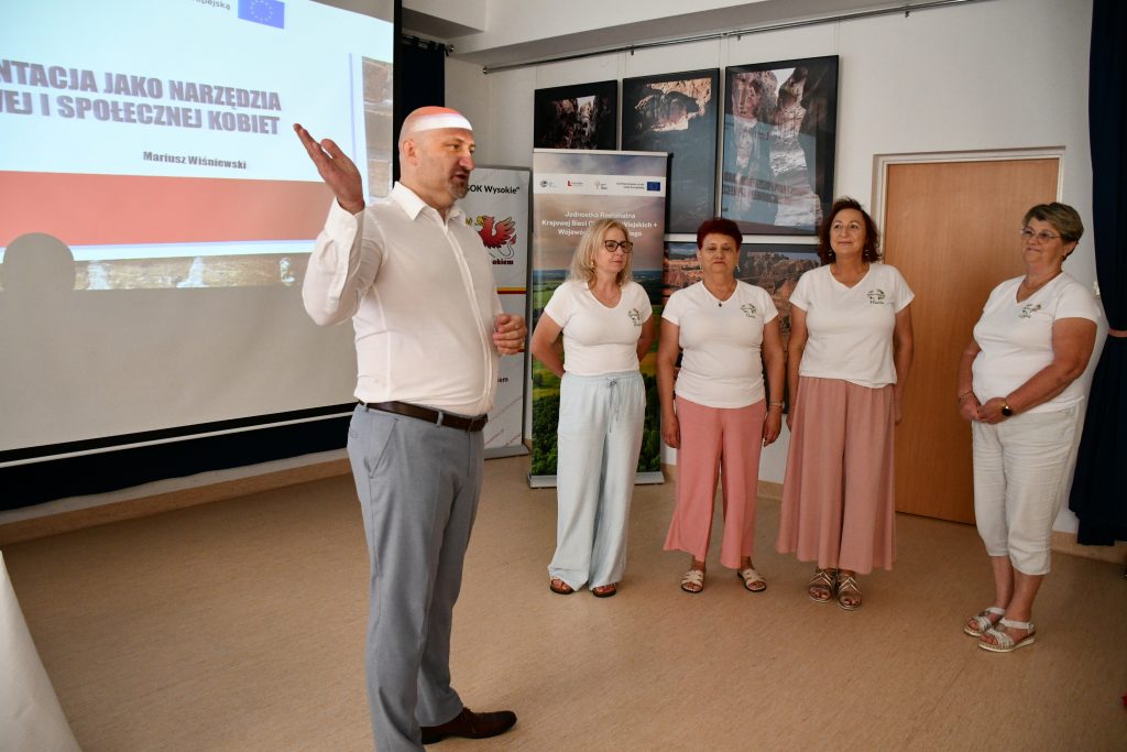 Prelegent prowadzący szkolenie z wizerunku i autoprezentacji oraz cztery uczestniczki skupione na omawianych zagadnieniach. W tle widoczny jest ekran z prezentacją multimedialną uzupełniającą temat wykładu.