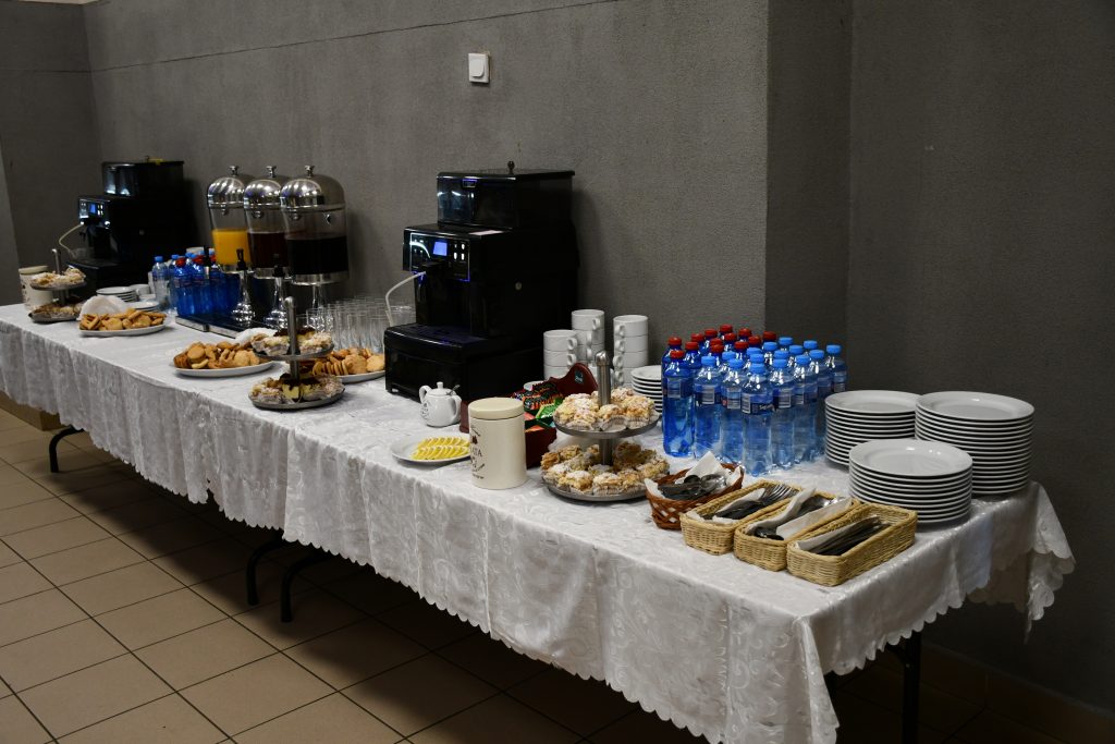 Stół cateringowy pokryty białym obrusem, na którym znajduje się poczęstunek dla uczestników-ciastka oraz napoje.